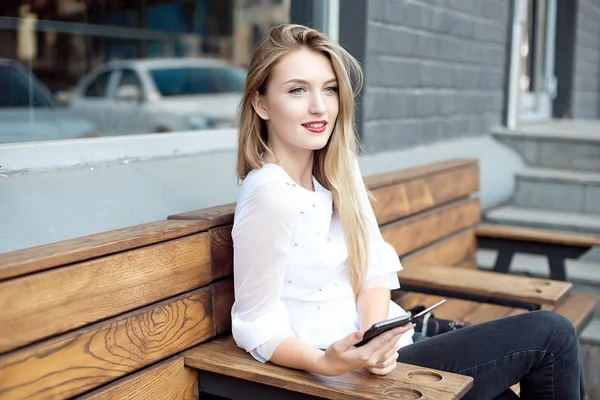 Attraktiv ung kvinna i Vintage jeans sitter på en stol i en utomhusrestaurang. Attraktiv blondin i en vit skjorta väntar på en beställning i sitt favoritkafé och njuter av naturen. — Stockfoto