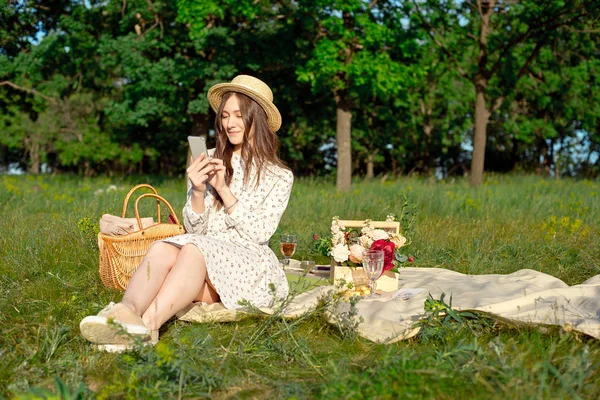 夏 - 牧草地でのピクニック。座っている女の子、スマートフォンで本を読んで、ピクニックバスケットとバゲットの近く、ワイン、グラス、ブドウ、ロール. — ストック写真
