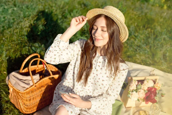 フィールドで日没ピクニックで麦わら帽子をかぶった若い美しい、黒髪の女の子。秋。バスケットとブーケで果物や飲み物をキャンプ. — ストック写真