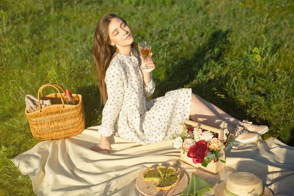 彼女の手にワインのグラスを保持する白い毛布の上に座って芝生の上にピクニックの若いブルネットは、夕日を楽しんで. — ストック写真