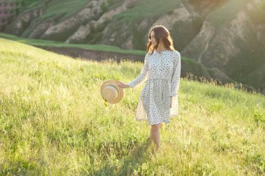 Elinde hasır şapkalı mutlu kadın, gökyüzünün gün batımı ışığı altında bir dağın kenarında yürürken, başarının, özgürlüğün ve parlak geleceğin tadını çıkarıyor..