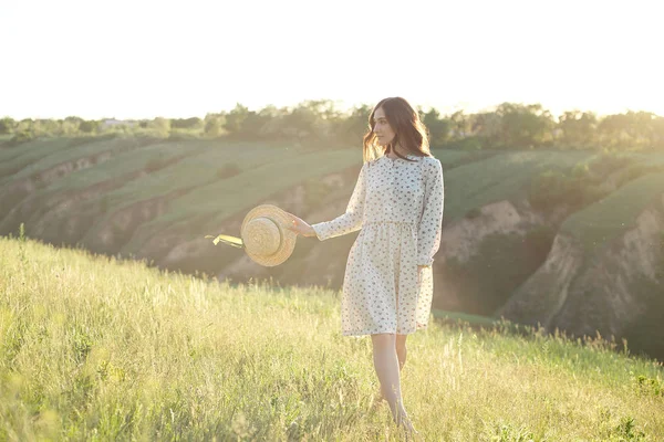 空の夕日の光の下で山の崖の端を歩き、成功、自由、明るい未来を楽しむ彼女の手に麦わら帽子を持つ幸せな女性. — ストック写真