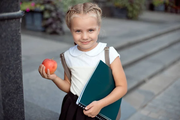 Felice scolaretta con i capelli biondi in uniforme scolastica con pranzo, libro siede vicino alla scuola . — Foto Stock