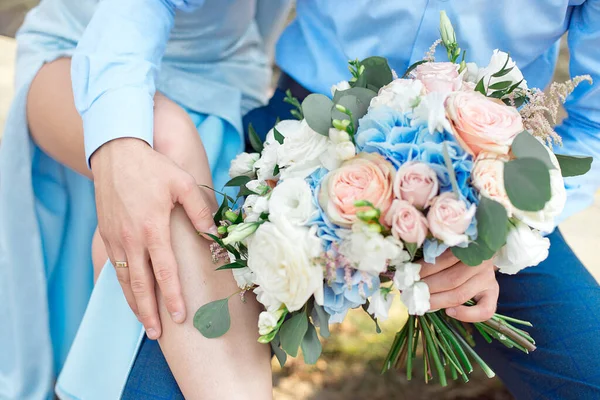 Primo piano della sposa e dello sposo, seduti su una panchina nel parco, lo sposo abbraccia la sposa, tenendo un bouquet nelle mani dell'aria aperta — Foto Stock