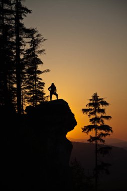 Renkli bir gün batımına karşı kayalık bir uçurumda poz veren genç bir kadının siluetini