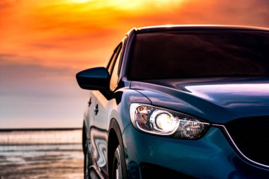 Mavi kompakt Suv araç spor ve modern tasarımı ile gün batımında sahilde denizin kenarında park etmiş. Çevre dostu teknoloji. İş başarı kavramı. Önden görünüm araba açık Far ışığı ile.