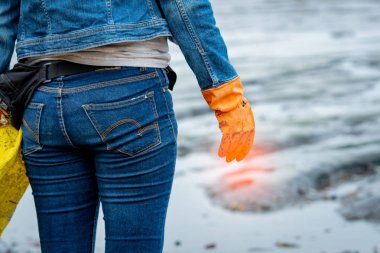 Gönüllü kot pantolon ve uzun kollu gömlek giymek ve sahilde çöp toplamak için turuncu lastik eldiven giymek. Plaj ortamı. Sahile temizlik kadın. Sahilde çöp toplama.