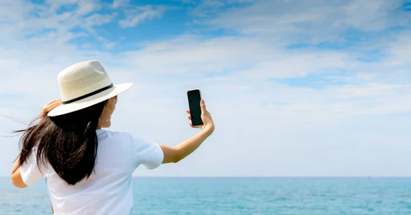 Młoda kobieta azjatyckich nosić kapelusz w stylu casual użytkowania smartfona biorąc selfie na molo. Letnie wakacje w tropikalnym raju plaży. Szczęśliwa dziewczyna podróży na wakacje. Kobieta cieszyć się i zrelaksować się życia. Letnie wibracje. — Zdjęcie stockowe