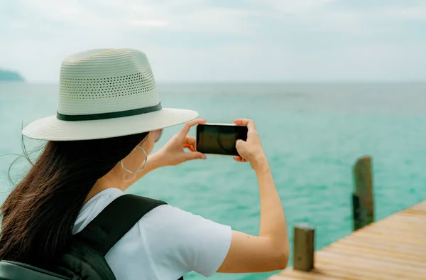 Młoda Azjatka backpacker kobieta nosić kapelusz użytkowania smartfona biorąc zdjęcie molo. Letnie wakacje w tropikalnym raju plaży. Hipster szczęśliwy dziewczyna podróży na wakacje. Kobieta cieszyć się i zrelaksować się życia. Letnie wibracje. — Zdjęcie stockowe