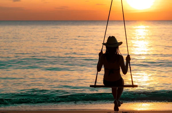 Siluett kvinna bära bikini och stråhatt swing gungor på stranden på sommarsemester vid solnedgången. Flicka i badkläder sitta på gungor och titta vackra solnedgången. Sommar vibbar. Kvinna resa ensam. — Stockfoto