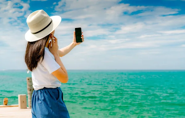 Młoda kobieta azjatyckich nosić kapelusz w stylu casual użytkowania smartfona biorąc selfie na molo. Letnie wakacje w tropikalnym raju plaży. Szczęśliwa dziewczyna podróży na wakacje. Kobieta cieszyć się i zrelaksować się życia. Letnie wibracje. — Zdjęcie stockowe