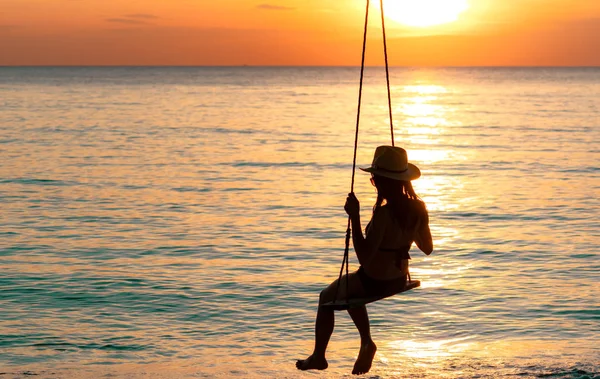 Siluett kvinna bära bikini och stråhatt swing gungor på stranden på sommarsemester vid solnedgången. Att njuta och avkopplande flicka på semester. Sommar vibbar. Kvinnan tittar på vackra solnedgång sky. — Stockfoto