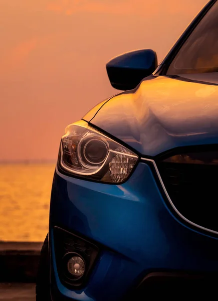 Vedere din față a mașinii SUV albastre cu design sportiv și modern, parcate pe șoseaua de beton lângă mare la apusul soarelui seara. Faruri de închidere lumina de masina albastra. Conceptul de tehnologie hibridă și electrică . — Fotografie, imagine de stoc