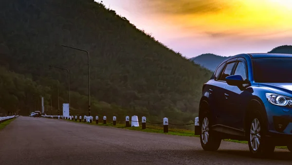 Azul SUV coche aparcado en la carretera de asfalto en frente de la montaña. Coche aparcado en el parque por la mañana temprano con el cielo del amanecer. Viaje por carretera. Plaza de aparcamiento. Industria automotriz. Turismo de naturaleza . — Foto de Stock