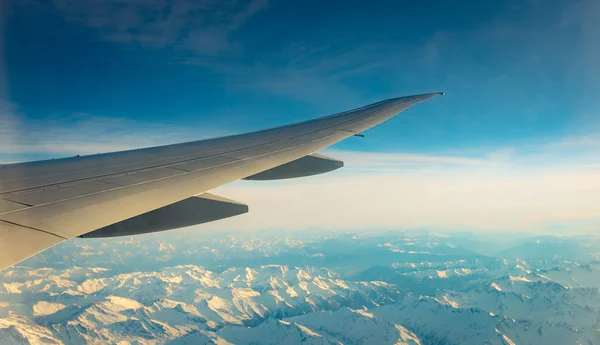 Wing of plane over mountain cover with white snow. Airplane flying on blue sky. Scenic view from airplane window. Commercial airline flight. Plane wing. Flight mechanics concept. International flight.