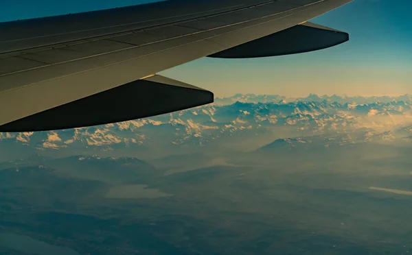 Wing of plane over mountain cover with white snow. Airplane flying on blue sky. Scenic view from airplane window. Commercial airline flight. Plane wing. Flight mechanics concept. International flight.