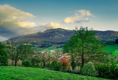 Tepenin tepesinde güzel bir manzara. Vadideki köy. Mavi gökyüzü ve beyaz bulutlar ile erken baharda ağaç. Kaya Dağı 'na güneş ışığı. Avrupa 'da ilkbahar sezonu. Tepede yeşil çim alan peyzaj. 