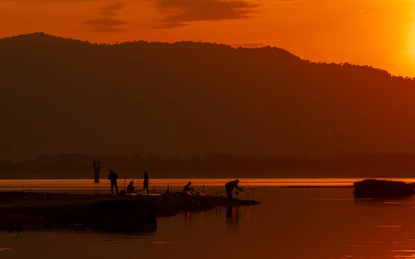 Vacker soluppgång himmel ovanför berget vid reservoaren. Folk fiskar med ett fiskespö på älven. Landskap av reservoar och berg med orange soluppgång himmel. Siluett liv på morgonen. — Stockfoto