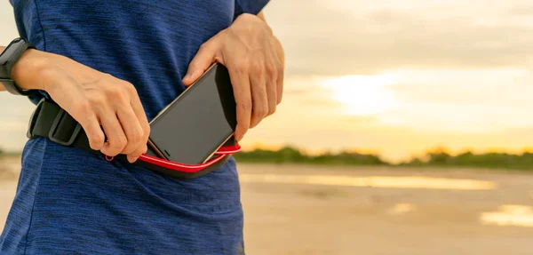 Mujer asiática joven mantener el teléfono inteligente en la bolsa de cintura antes de ejecutar ejercicio cardiovascular por la mañana. Entrenamiento al aire libre. Corredor y dispositivo portátil banda inteligente. Gadget para un estilo de vida saludable. Moda jogging . — Foto de Stock