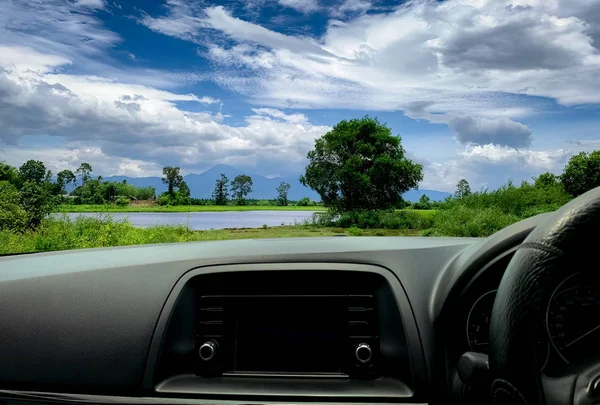 Beautiful landscape view from inside car. Steering wheel and dashboard of car interior. Road trip travel with scenic view of mountain, lake, and forest. Blue sky and white fluffy clouds. Vacation time.