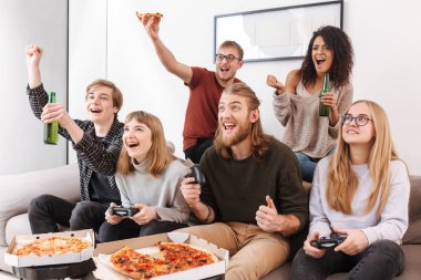 Kanepede oturan ve birlikte video oyunları oynarken vakit neşeli arkadaş grubu. Pizza yiyip evde bira içmeyi gençlerin portresi