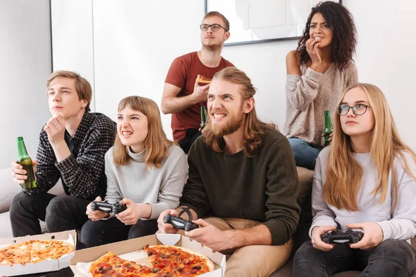 Group of friends sitting on sofa and spending time together while playing video games,eating pizza and drinking beer at home. Portrait of people showing different emotions while playing games