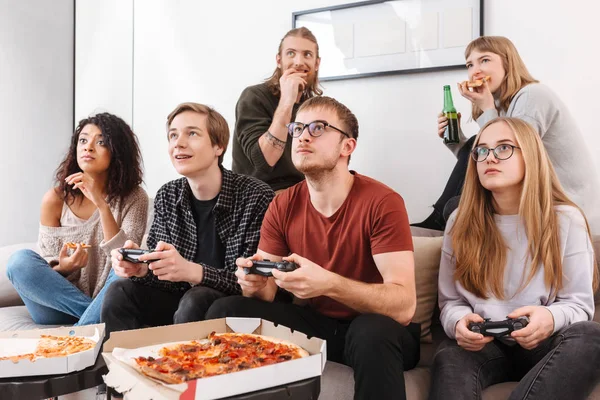 Group of thoughtful friends sitting on sofa and spending time together while playing video games,eating pizza and drinking beer at home