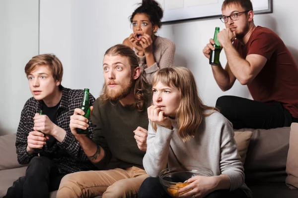 Group of friends sitting on sofa and intently watching movie together with chips and beer at home. Group of young people showing defferents emotions while watching film