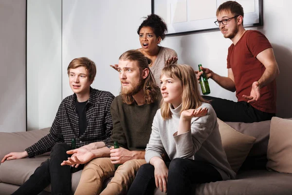 Group of amazed friends sitting on sofa and watching movie together with  beer at home. Group of young people watching film without interest