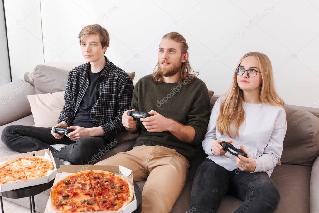 Group of friends sitting on sofa and playing video games together at home. Portrait of two boys and girl eating pizza and playing games isolated