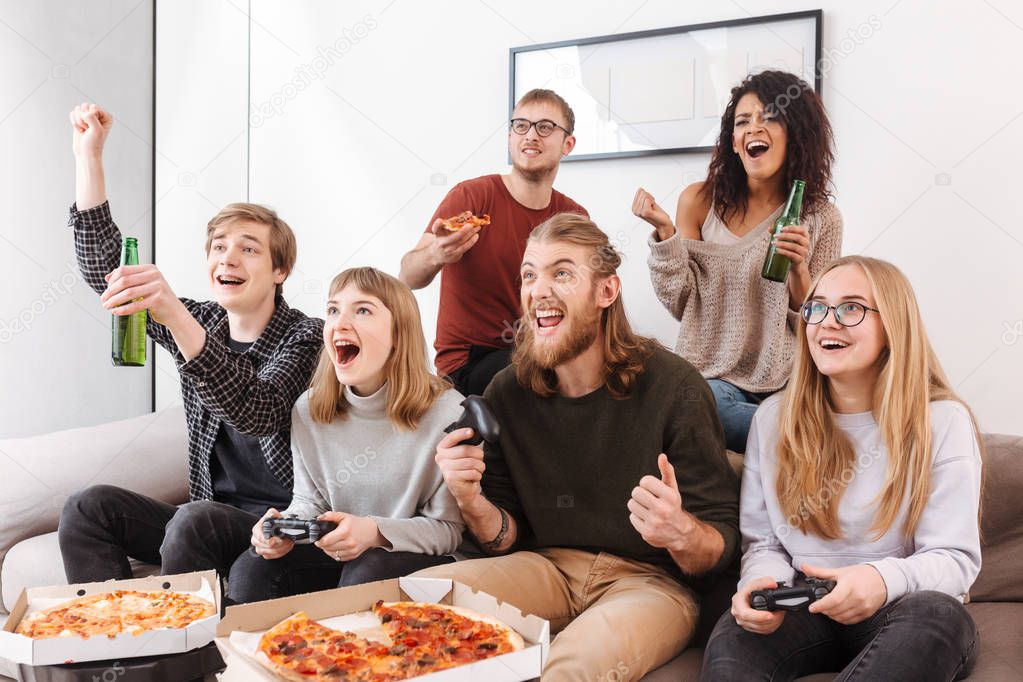 Group of joyful friends sitting on sofa and screaming yeah while spending time together and playing video games. Portrait of young people eating pizza and drinking beer at home