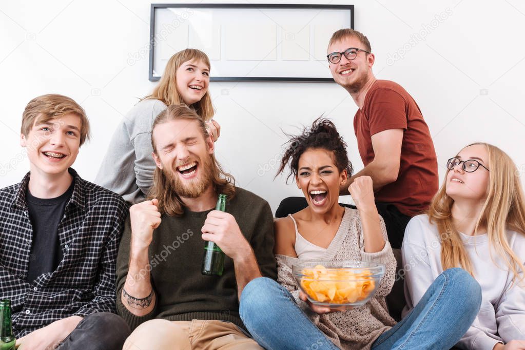 Group of joyful friends sitting on sofa and watching film together with chips and beer at home