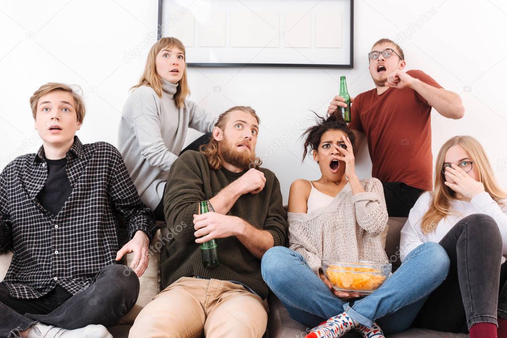 Close up photo of scared friends sitting on sofa and watching horror movie together with chips and beer at home