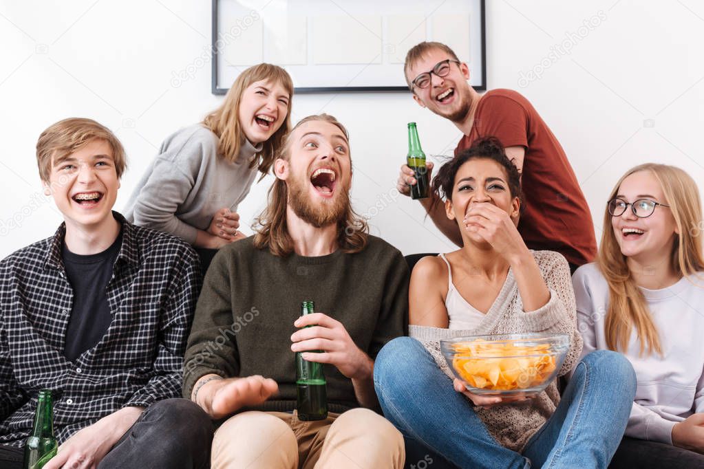 Group of happy laughing friends sitting on sofa and watching film together with chips and beer at home