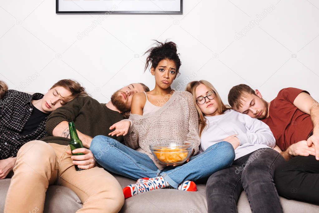 Portrait of young pretty lady sitting on sofa with chips and sadly looking in camera while her friends sleeping near at home
