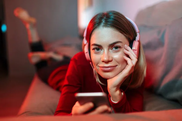 Portrait of pretty smiling lady in headphones lying on sofa with cellphone in hands and dreamily looking in camera at home