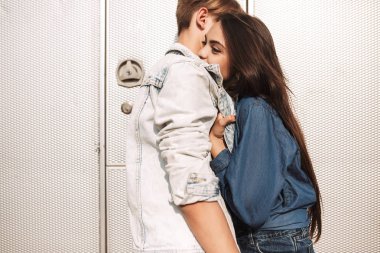 Ayakta ve başka bir arka plan üzerinde gri römork ile kucaklayan genç güzel çift portresi. Güzel Bayan mutlu erkek arkadaşıyla sarılma
