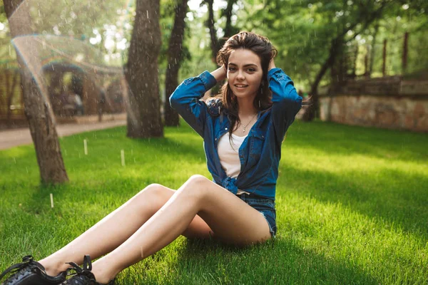 ショート パンツやデニム シャツ公園で雨の下で芝生の上に座って喜んでカメラを探している若い美しい女性 — ストック写真