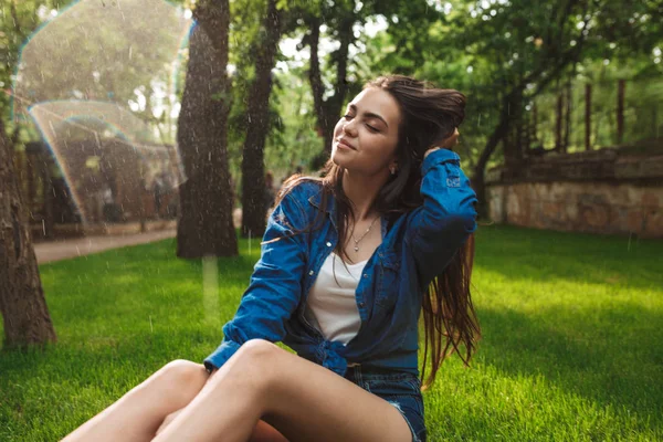 漂亮微笑的女士在短裤和牛仔衬衫的肖像朦胧关闭她的眼睛 而坐在草地下雨在公园 — 图库照片