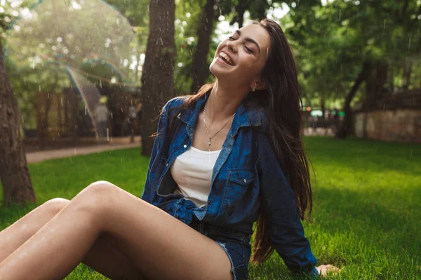 美丽微笑的女士在短裤和牛仔衬衫朦胧地闭上眼睛 而坐在草地下雨在公园 — 图库照片