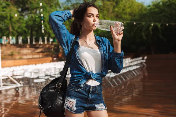 Portrait of young lady in denim shirt and shorts standing with black backpack and drinking water while thoughtfully looking aside under rain in park