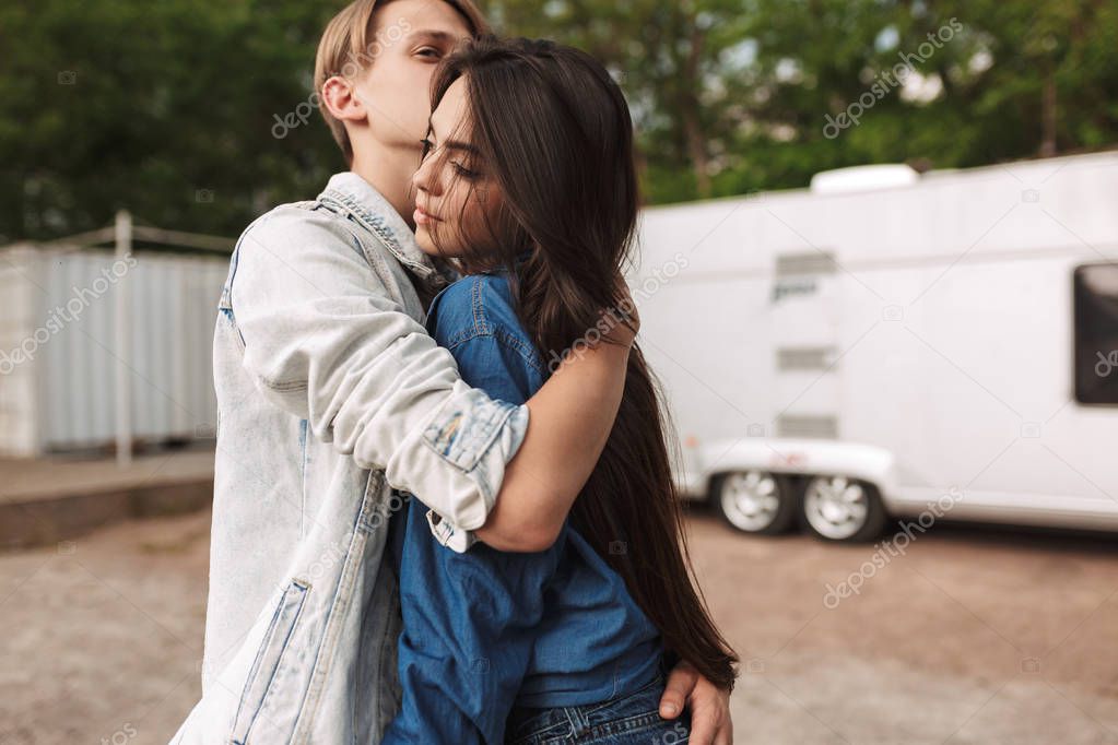 Portrait of young beautiful couple standing and embracing one another while spending time together in park with gray trailer on background