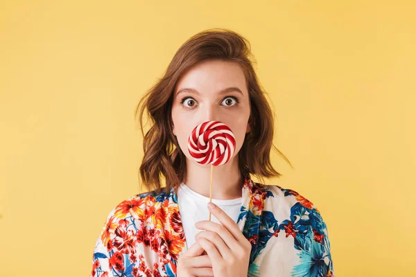 Portrait of young lady in colorful shirt standing and covering her lips with lollipop candy while amazedly looking in camera on over pink background