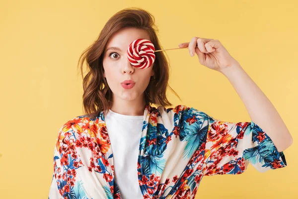 Portrait of beautiful lady in colorful shirt standing and covering her eye with lollipop candy while amazedly looking in camera on over pink background