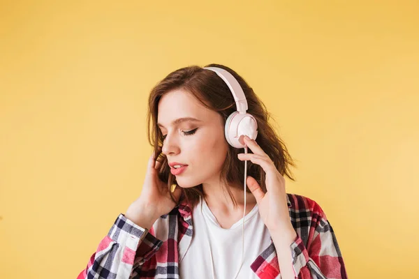 Portrait of beautiful pensive lady in colorful shirt standing and listening music in headphones on over pink background