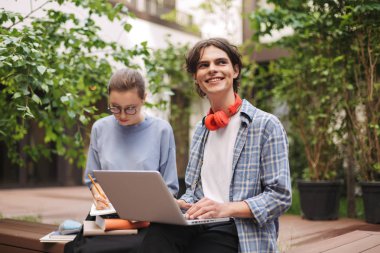 Dizüstü bilgisayar ile bankta oturmuş ve mutlu bir kenara Üniversitesi avluda arıyorum genç gülümseyen adam. Açık havada birlikte okuyan öğrenciler