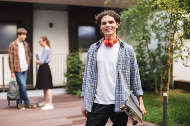 Neşeli çocuk ayakta ve mutlu bir şekilde fotoğraf makinesindeki Üniversitesi avluda öğrenciler ile arka plan üzerinde bakarken kaykay tutan kırmızı kulaklıklar ile