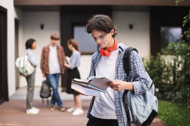 Sırt çantası ile ayakta ve arka plan öğrencilerle Üniversitesi avluda kitap okuma kırmızı kulaklık ile genç adam portresi