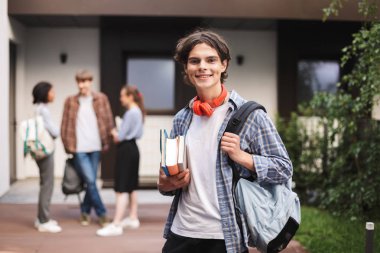 Neşeli çocuk kırmızı kulaklıklarla sırt çantası ve el ve mutlu bir şekilde fotoğraf makinesindeki Üniversitesi avluda öğrencilerle arka plan üzerinde seyir defterleri ile ayakta