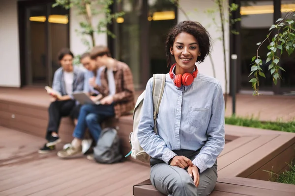 Smiling lady in shirt sitting on bench with red headphones and backpack in courtyard of university. Beautiful girl sitting and happily looking in camera with students on background